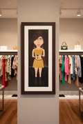 Framed artwork of a woman in a store setting with clothing racks and bags in the background