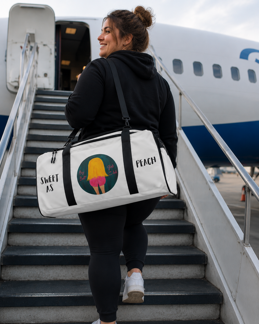 Person carrying a white duffel bag with graphics and text, walking up airplane stairs.
