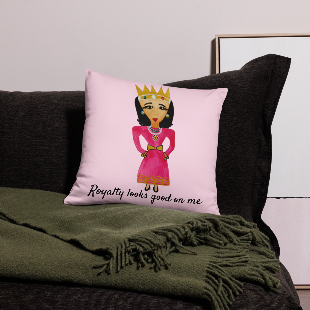 A square pink throw pillow with a cartoon illustration of a queen in a pink dress and crown, sitting on a dark gray sofa with an olive green throw blanket.