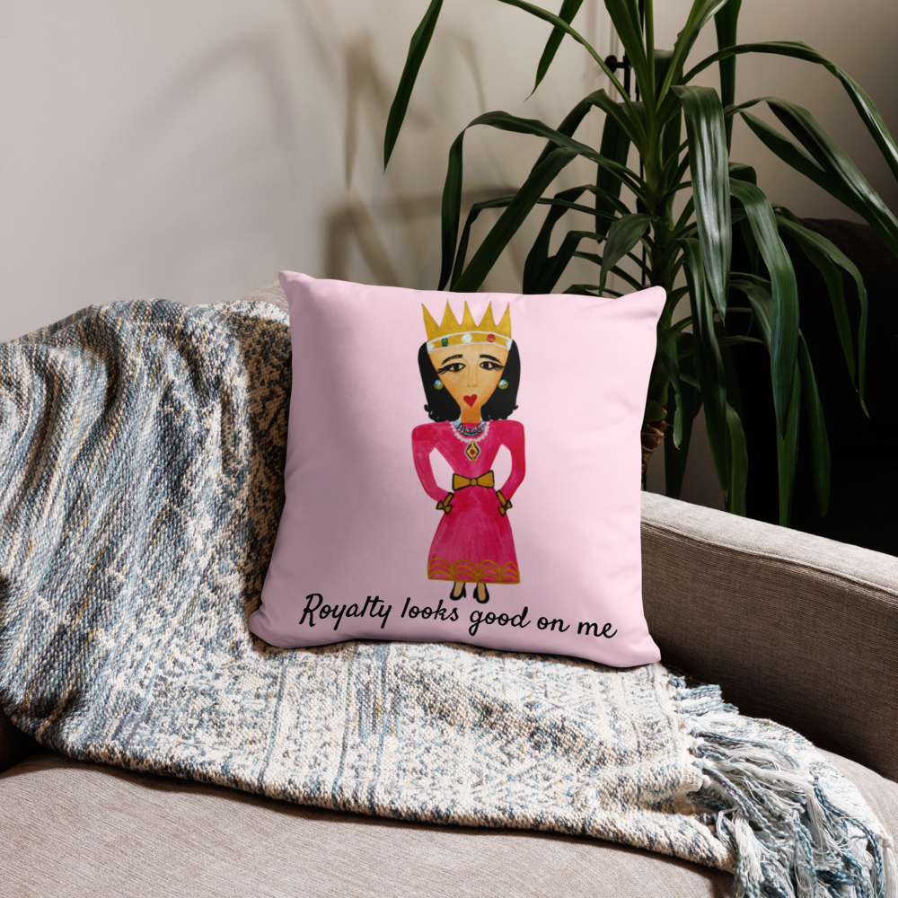 A square pink throw pillow with a cartoon illustration of a queen in a pink dress and gold crown, sitting on a beige sofa next to a blue-gray patterned throw blanket and a green houseplant.