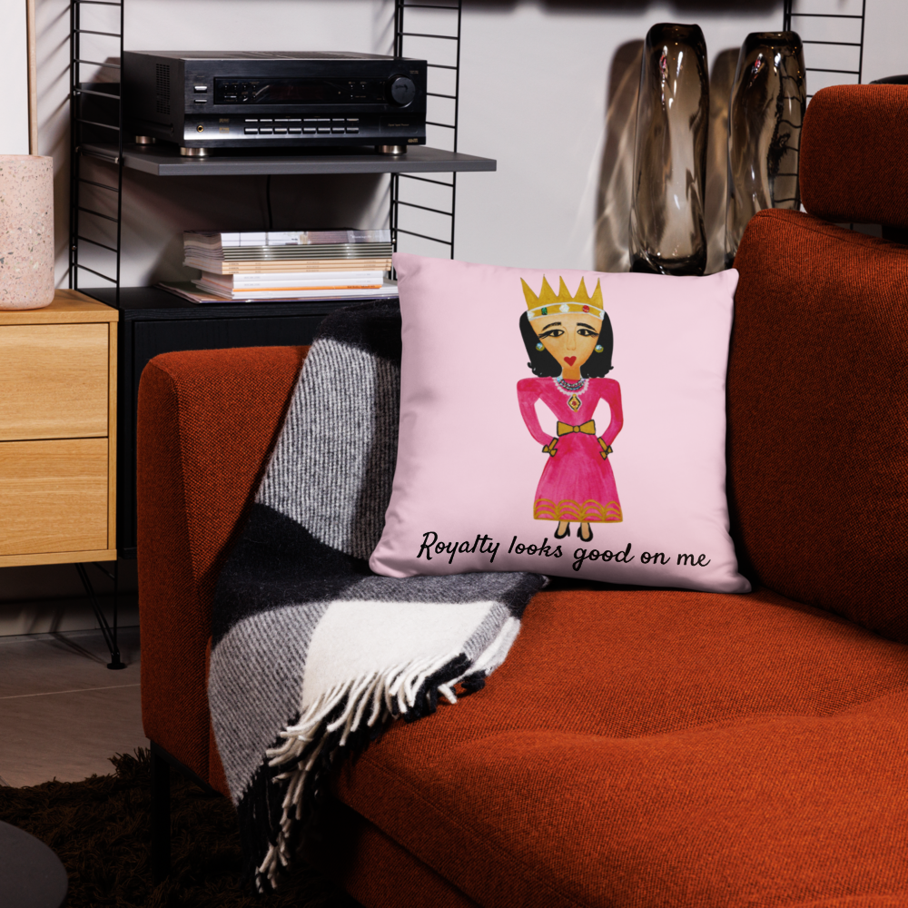 A square throw pillow with a pink background and a cartoon illustration of a queen in a pink dress and gold crown, sitting on an orange-brown couch next to a black-and-white plaid throw blanket.