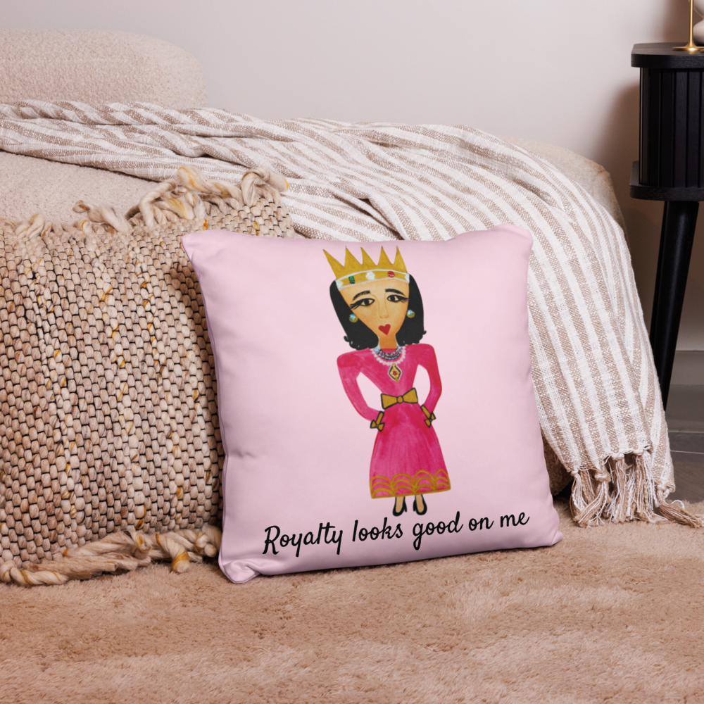 A square pink throw pillow with a cartoon illustration of a crowned queen in a pink dress and the text 'Royalty looks good on me', styled on a beige rug next to a textured knit blanket and striped throw.