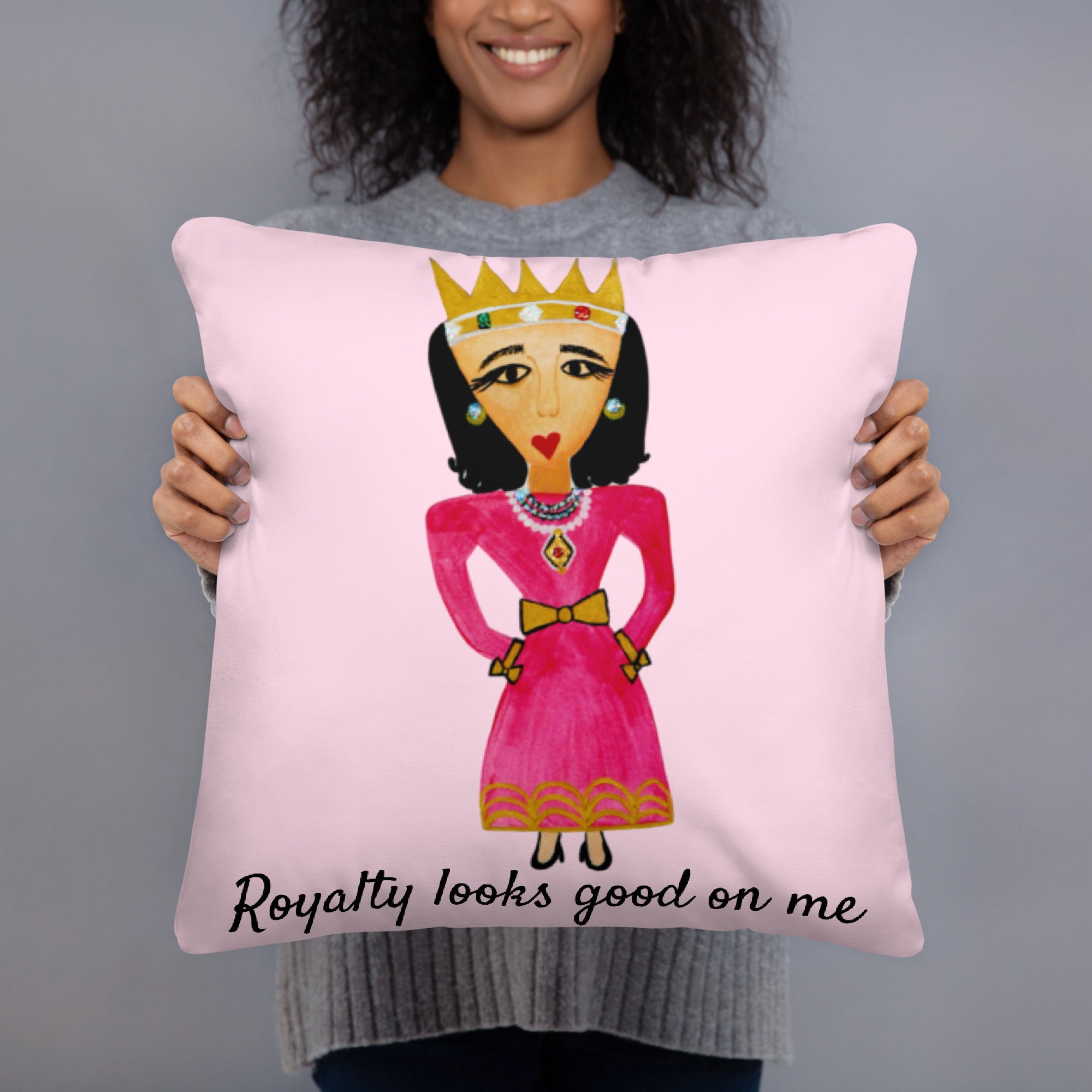 A person holding a square pink throw pillow featuring a stylized illustration of a crowned queen in a pink dress with the text 'Royalty looks good on me' at the bottom, against a plain gray background.