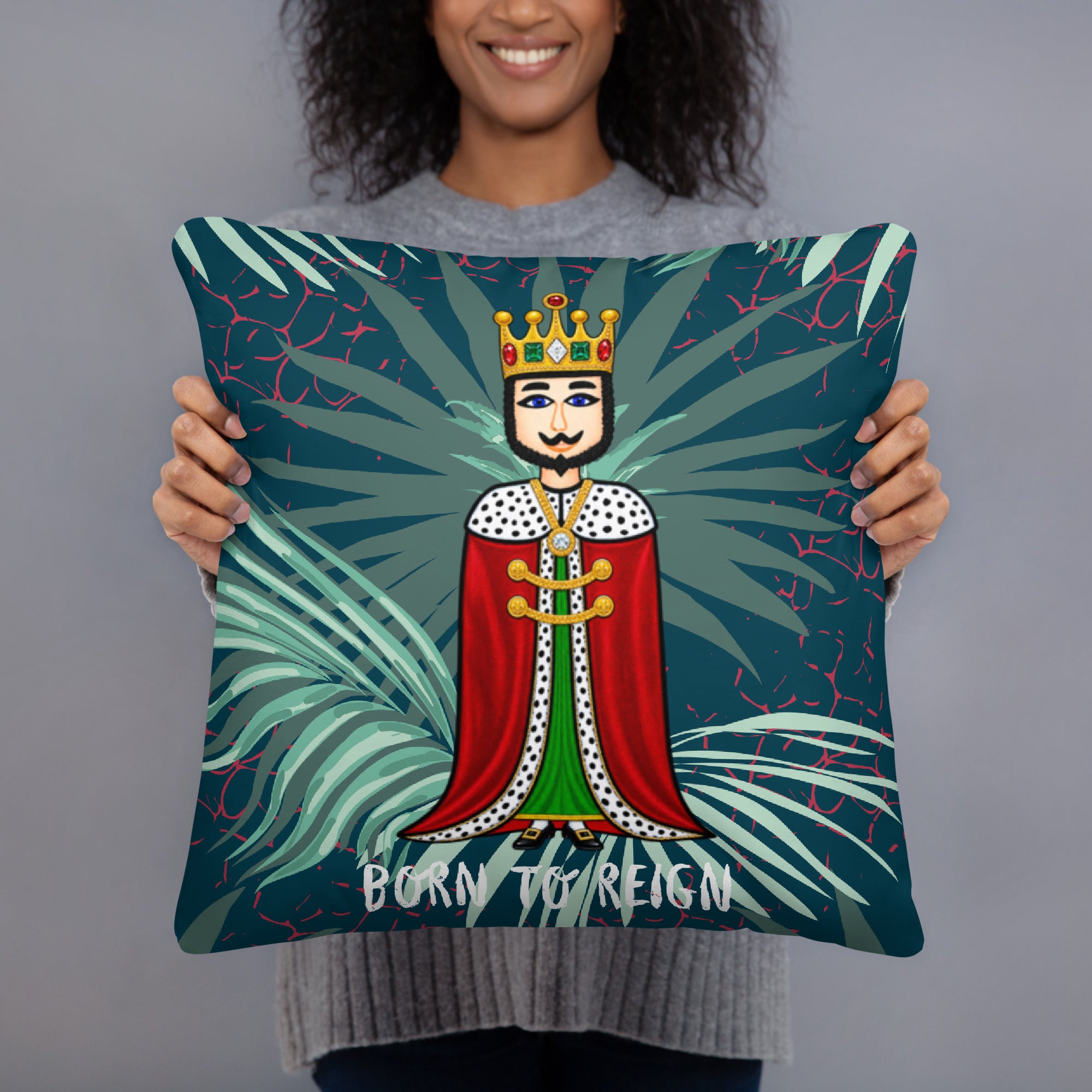 A person holding a square decorative throw pillow featuring a stylized cartoon king with a crown and red cape on a dark teal background with palm leaves and the text 'BORN TO REIGN'.