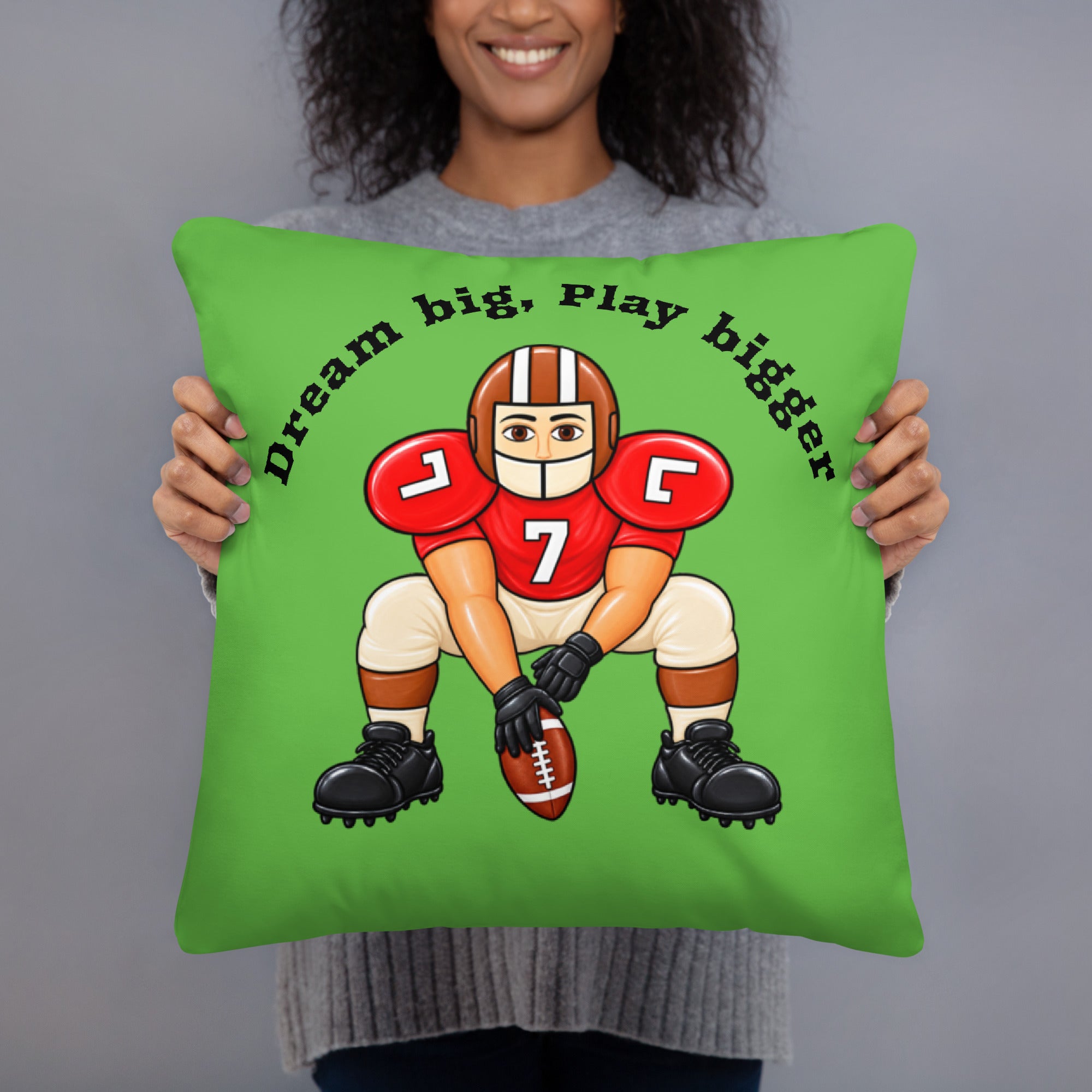 A person holding a bright green square throw pillow with a cartoon football player graphic and the text 'Dream big, Play bigger' on a plain gray studio background.
