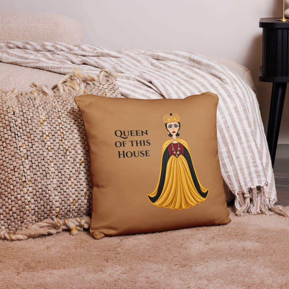 A square tan throw pillow with a printed graphic of a queen in a yellow and black gown and the text 'QUEEN OF THIS HOUSE', styled on a beige sofa with a striped blanket and textured knit throw.