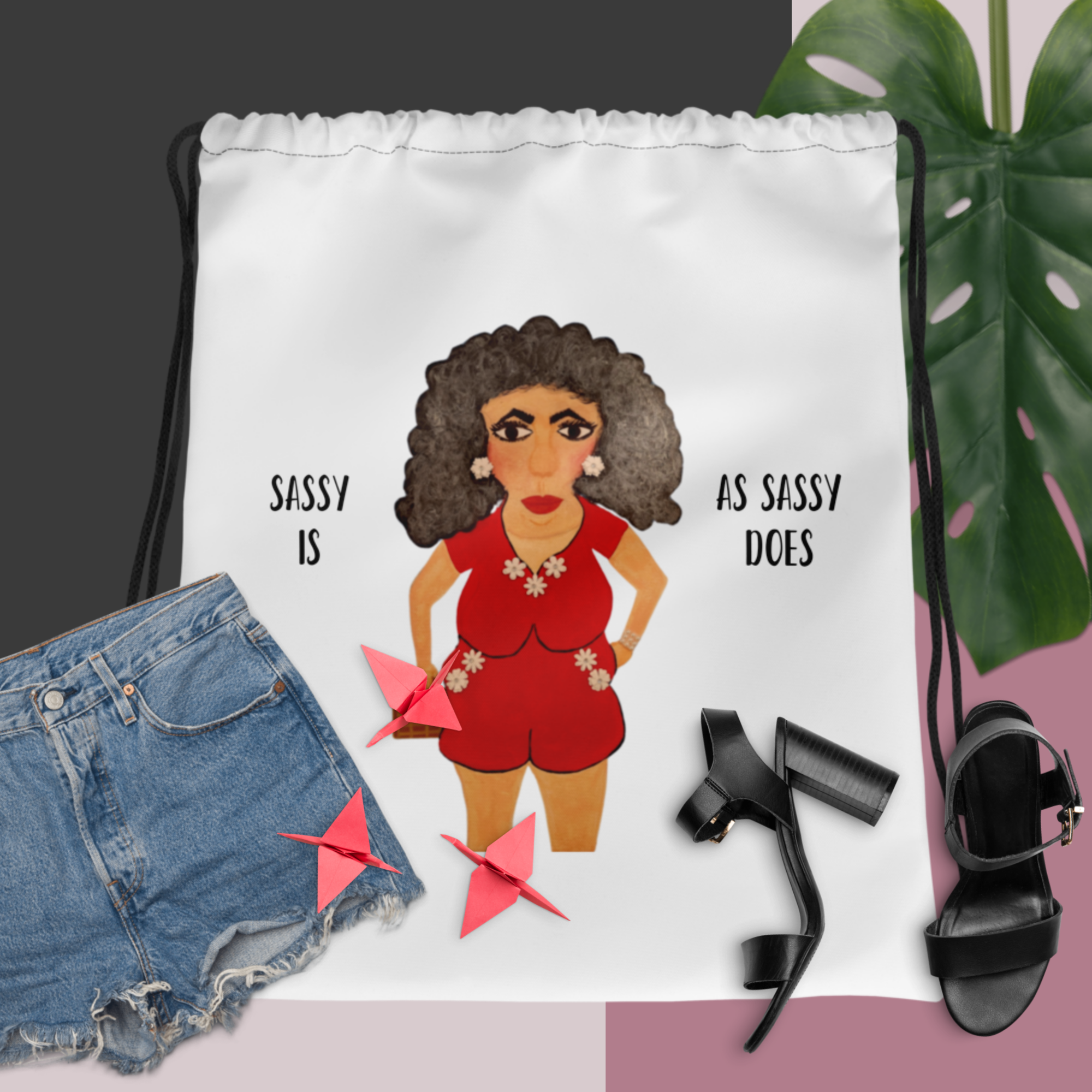 Drawstring bag with a graphic of a woman in a red outfit and text, surrounded by denim shorts, black high heels, and pink shoes.