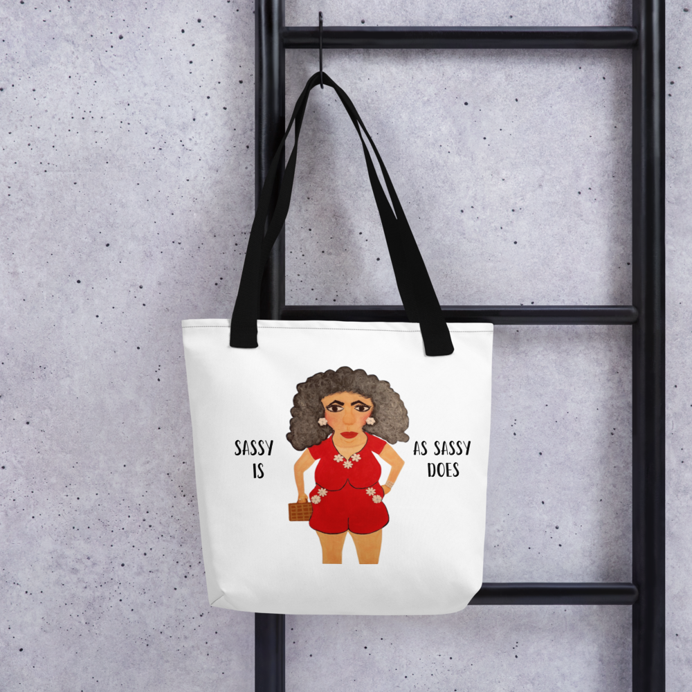 Tote bag with a graphic of a woman in a red outfit and text 'Sassy is as sassy does' hanging on a black metal rack against a gray concrete wall.