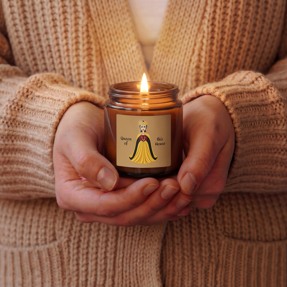 A person in a beige knit cardigan holds a lit amber glass jar candle with a cream label featuring a cartoon queen in a yellow dress and the text 'Queen of this house'.