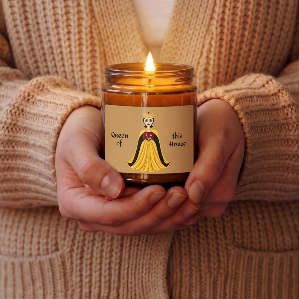 A person in a beige knit sweater holds a lit candle in a glass jar. The candle label features an illustration of a queen in a yellow gown with the text 'Queen of this House'.