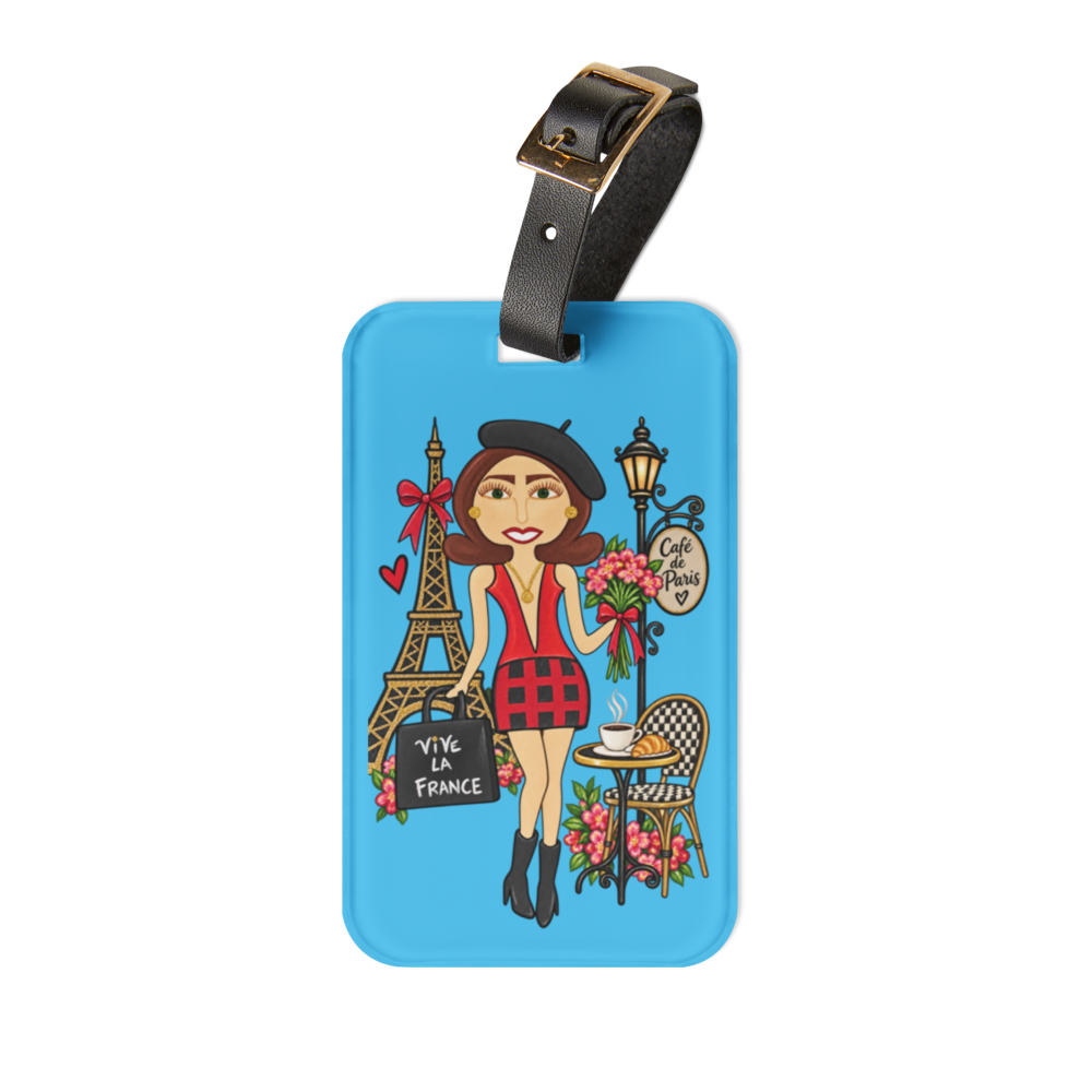 A rectangular blue luggage tag with rounded corners, featuring a colorful cartoon illustration of a woman in Paris next to the Eiffel Tower, a street lamp, flowers, and a sign reading 'Vive La France'. The tag has a black strap with a gold-tone buckle at the top.