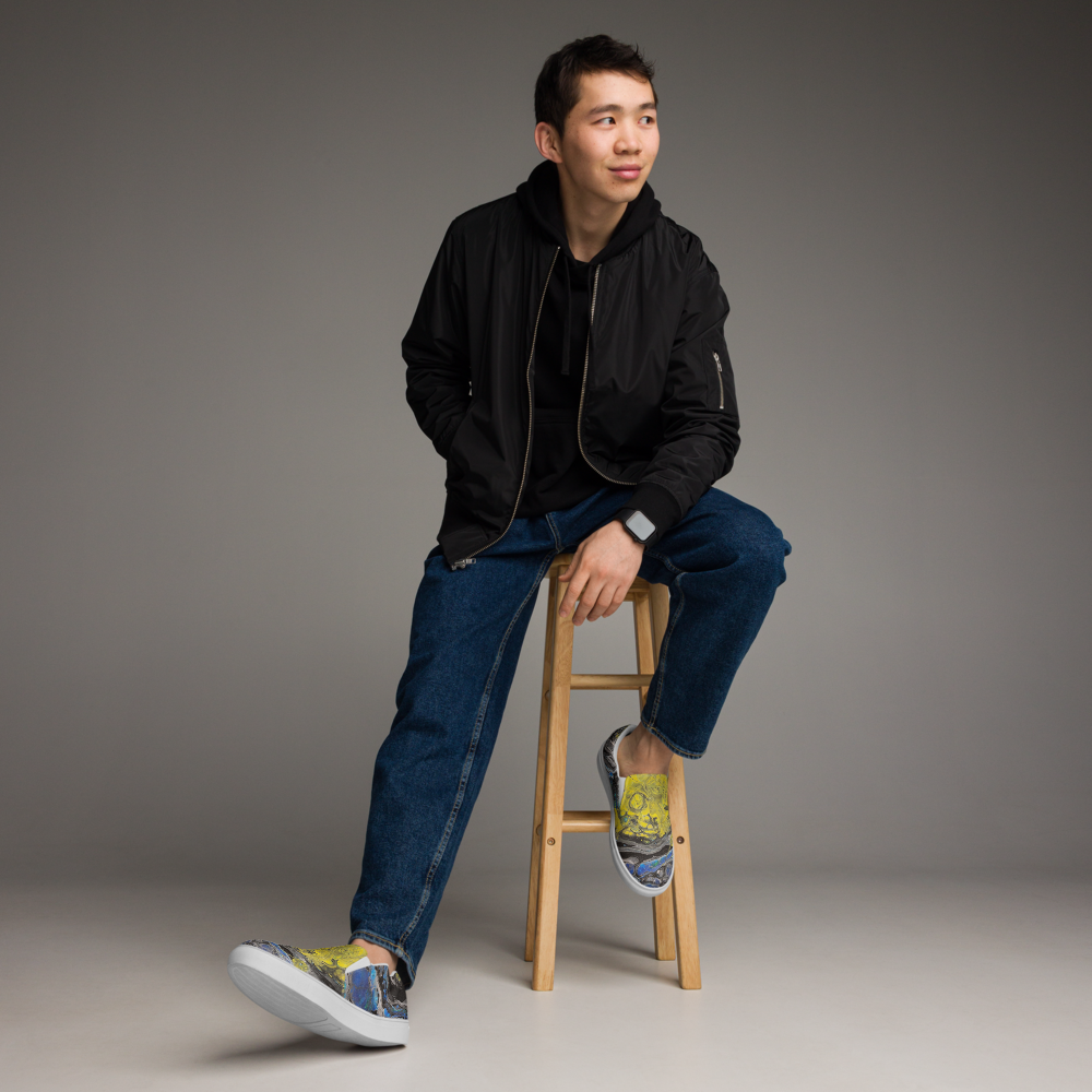 Man sitting on a wooden stool wearing a black jacket, blue jeans, and patterned shoes against a gray background