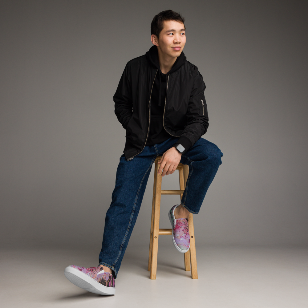 Man sitting on a wooden stool wearing a black jacket, blue jeans, and gray sneakers against a gray background