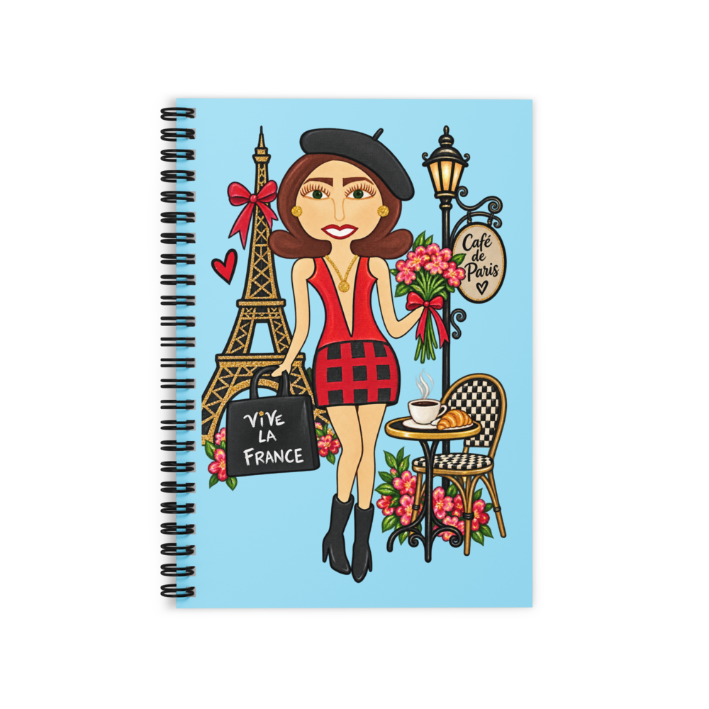 A spiral-bound notebook with a light blue cover featuring a cartoon illustration of a woman in Paris attire, including the Eiffel Tower, a flower bouquet, a street lamp reading 'Café de Paris,' and a black bag that says 'VIVE LA FRANCE.'.