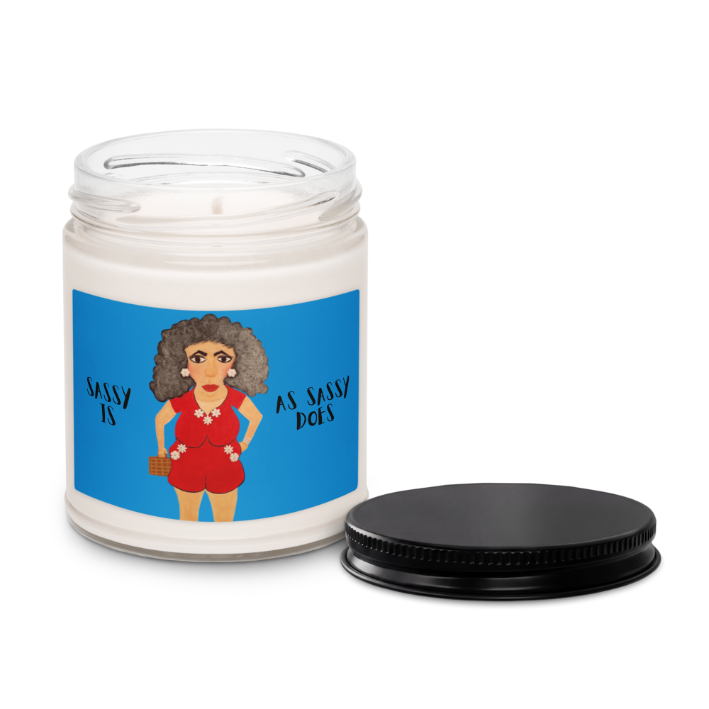 A white wax candle in a clear glass jar with a blue illustrated label showing a woman in a red outfit and the text 'SASSY IS AS SASSY DOES', shown with a black metal lid placed beside it on a white background.