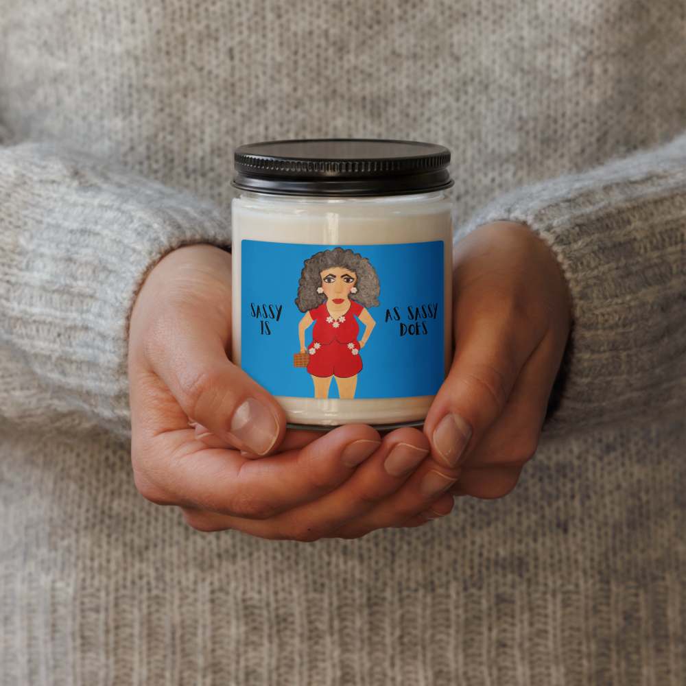 A person's hands holding a small glass jar candle with a black lid and a colorful label featuring an illustration of a woman with curly hair and the text 'MANY IS AS MANY DOES'. The background shows a person wearing a cozy gray knit sweater.