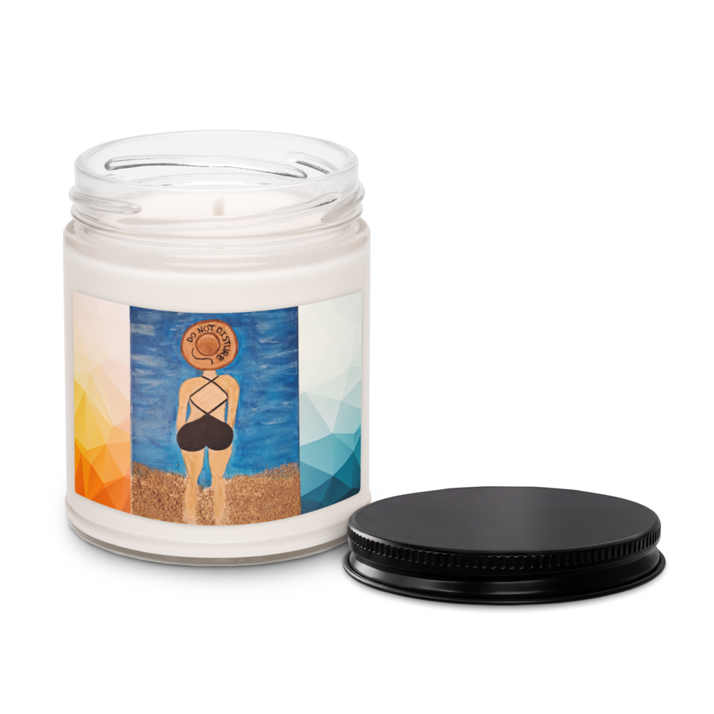 A white wax candle in a clear glass jar with a colorful label showing a beach scene, with a black metal lid placed beside it on a white background.