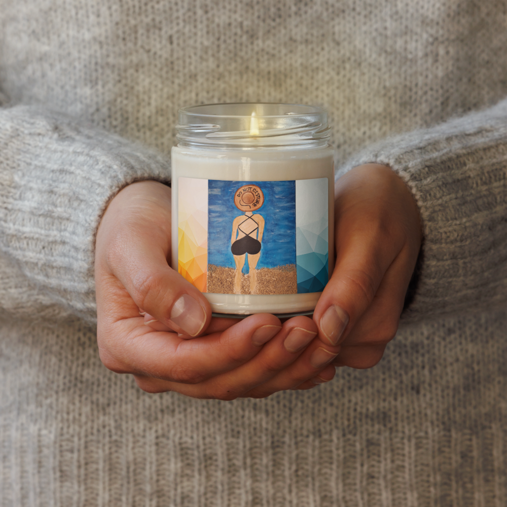 A person in a beige knit sweater holds a lit glass jar candle. The candle has a white wax body and a colorful label featuring an illustration of a person standing on a beach against a blue sky.