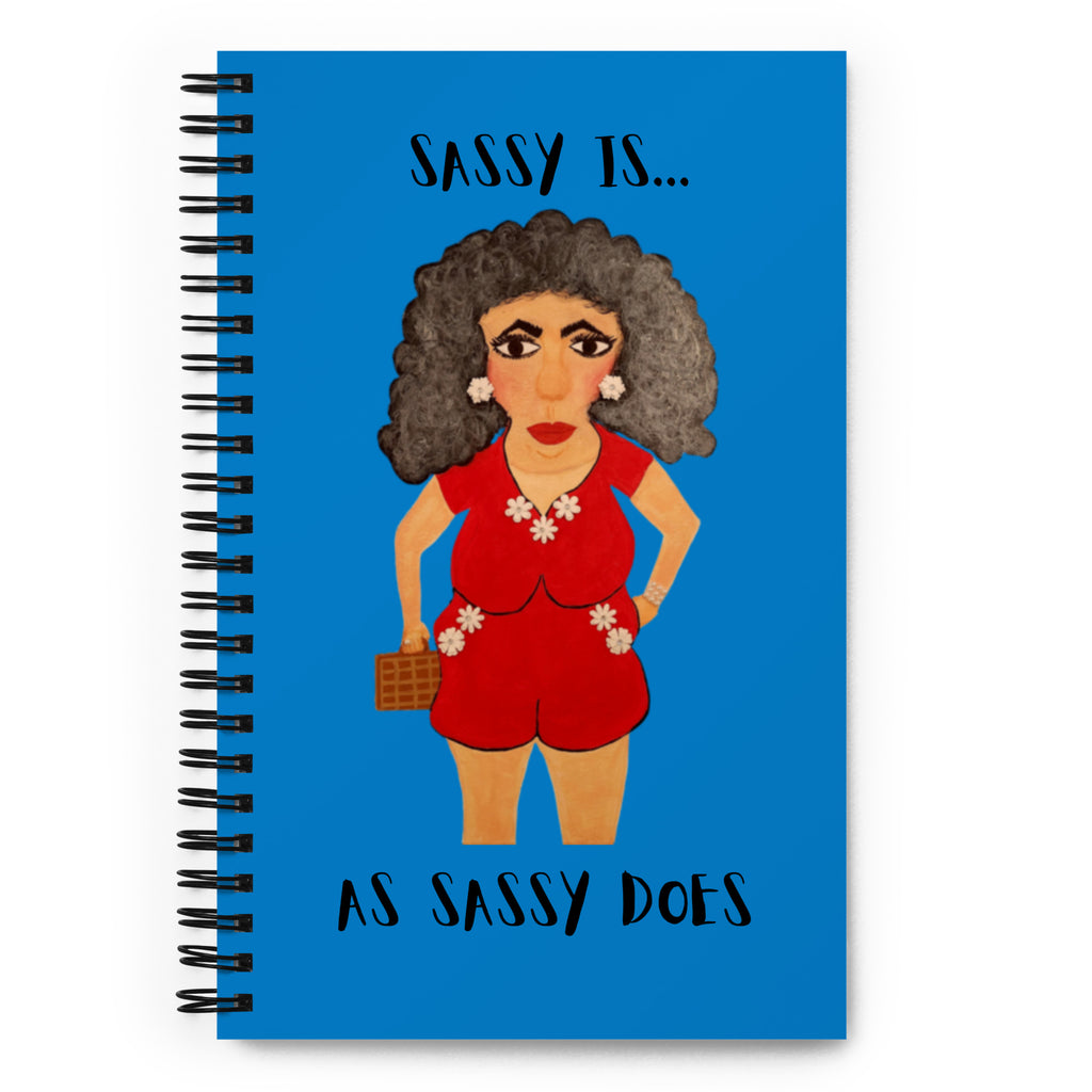A spiral-bound notebook with a bright blue cover featuring an illustrated woman in a red outfit and the text 'SASSY IS.