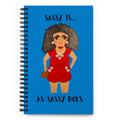 A spiral-bound notebook with a bright blue cover featuring an illustrated woman in a red outfit and the text 'SASSY IS.