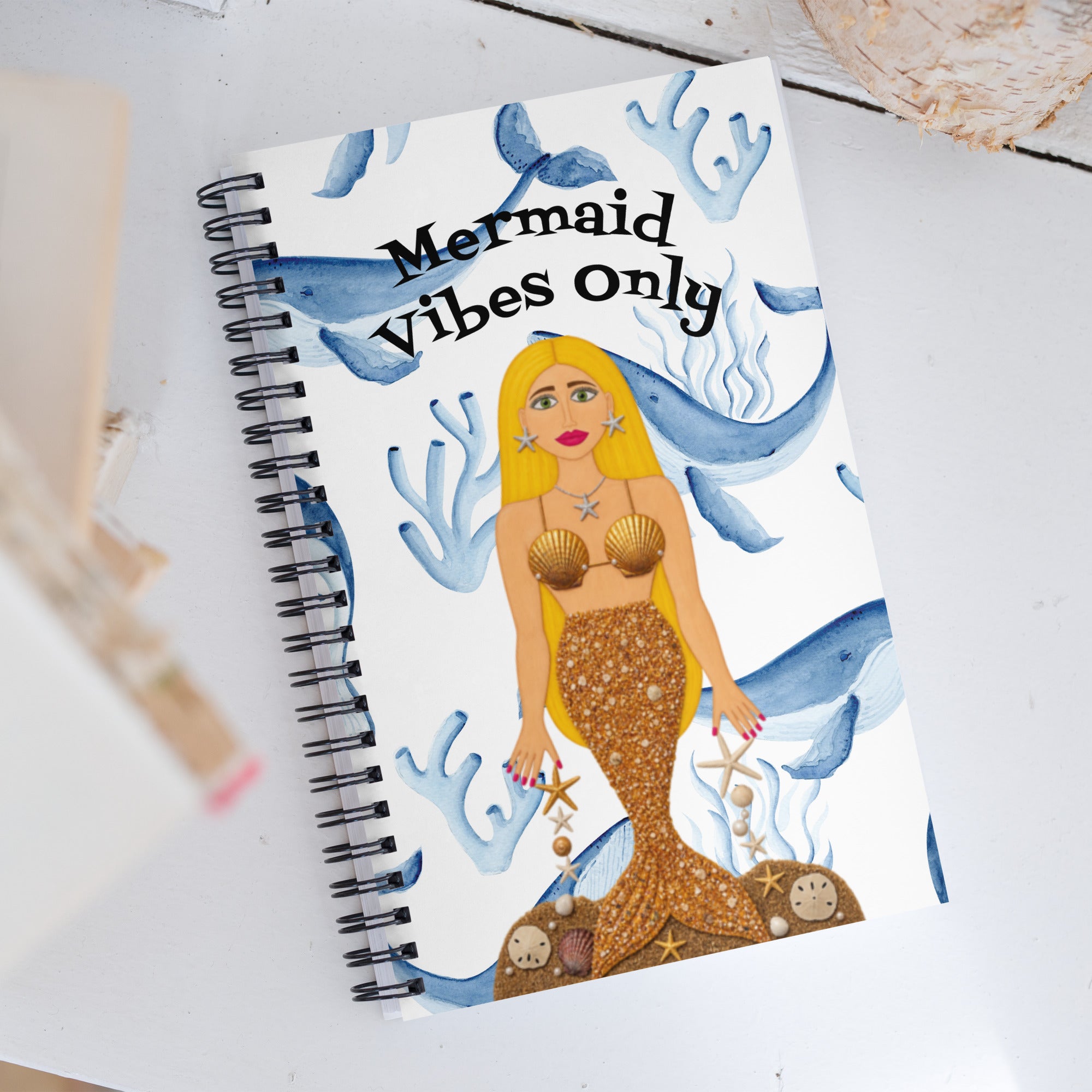 A spiral-bound notebook with a mermaid illustration and the text 'Mermaid Vibes Only' on a white cover, photographed on a light tabletop with a few blurred props nearby.