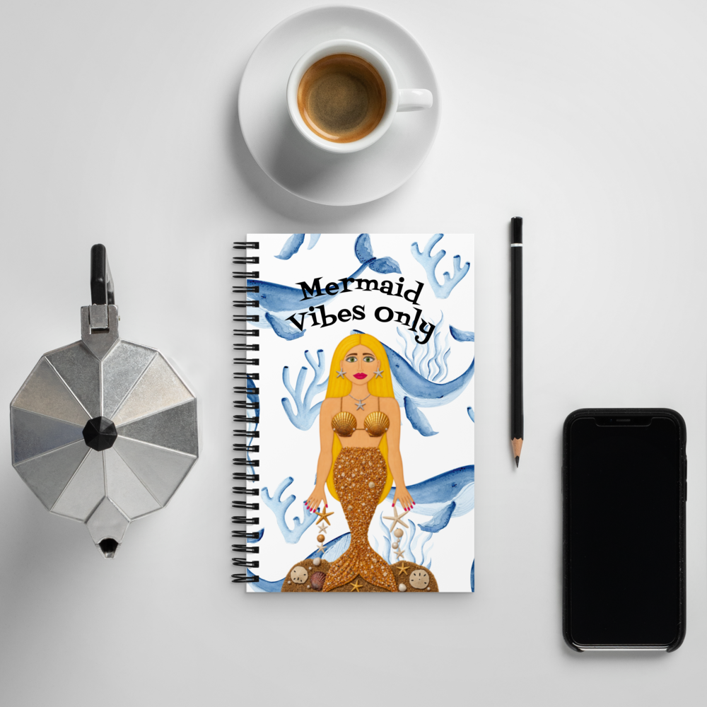 A spiral-bound notebook with a mermaid illustration and the text 'Mermaid vibes only' is arranged in a flat lay on a light gray surface, surrounded by a cup of coffee, a moka pot, a pencil, and a smartphone.