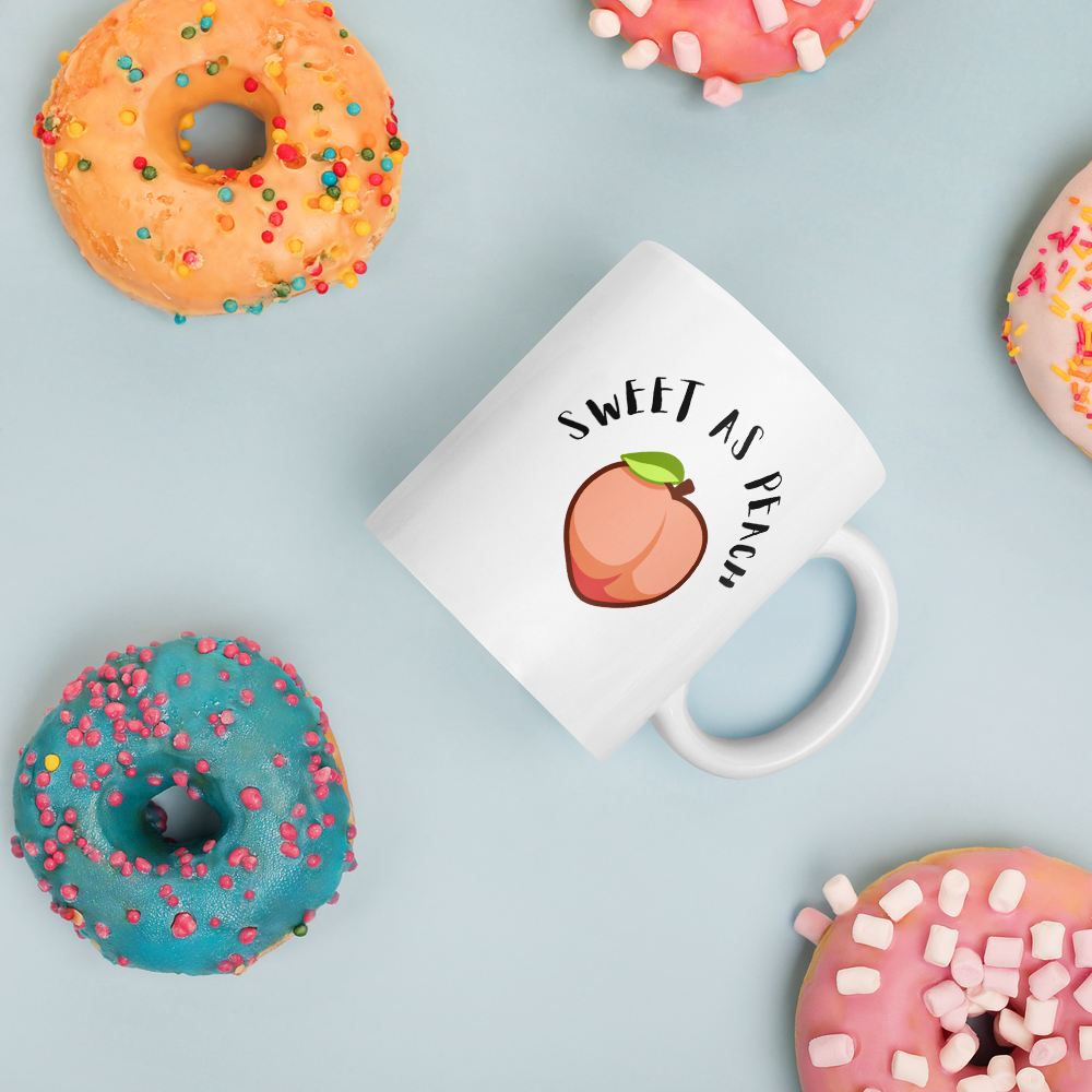 White mug with peach illustration and 'Sweet as Peach' text on a light blue background with colorful donuts.
