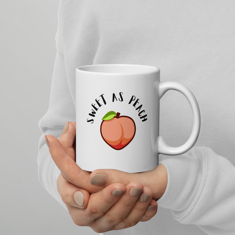 White mug with 'Sweet as Peach' design held by a person against a gray background
