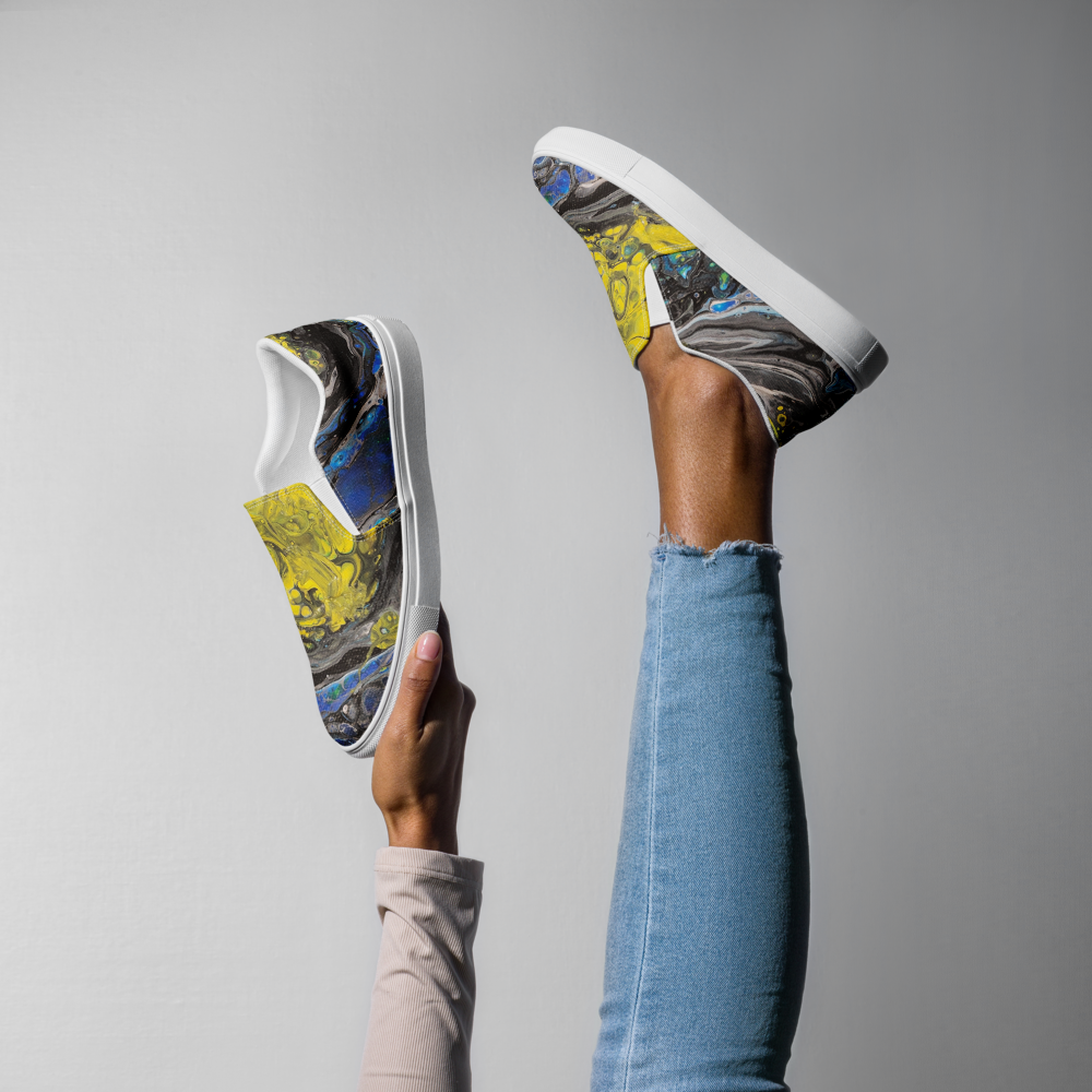 Colorful slip-on sneakers with abstract design held by a person wearing blue jeans against a gray background